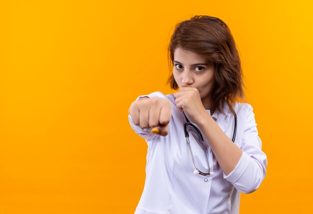 Médico joven en bata blanca con estetoscopio golpeando el puño apuntando a la cámara con él