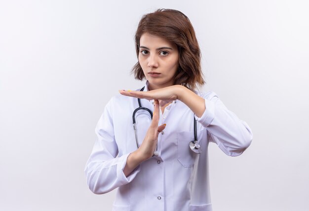 Médico joven en bata blanca con estetoscopio mirando cansado haciendo gesto de tiempo con las manos