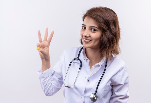 Médico joven en bata blanca con estetoscopio mostrando y apuntando hacia arriba con los dedos número tres sonriendo de pie sobre la pared blanca