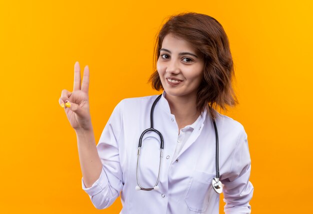 Médico joven en bata blanca con estetoscopio mostrando el signo de la victoria sonriendo de pie sobre la pared blanca