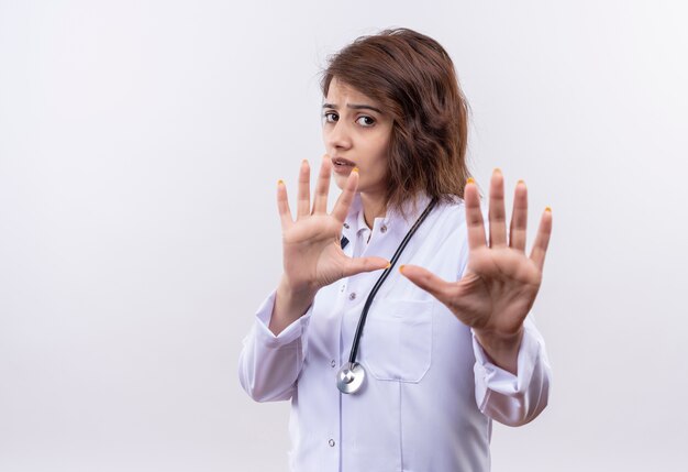 Médico joven en bata blanca con estetoscopio de pie con las manos abiertas haciendo señal de stop con aspecto preocupado sobre fondo blanco.