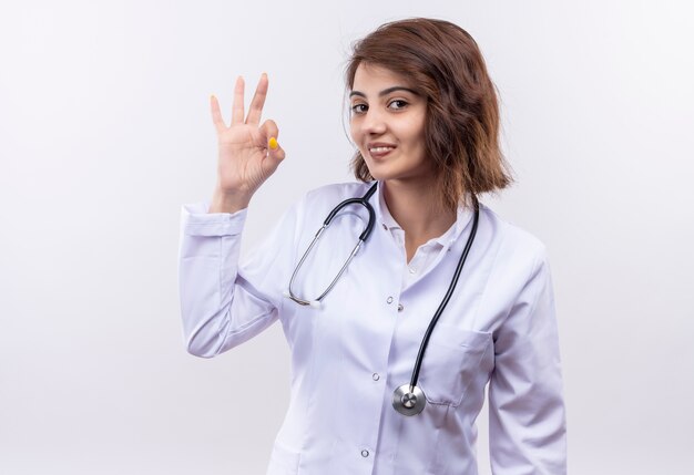 Médico joven en bata blanca con estetoscopio sonriendo confiado haciendo bien firmar de pie sobre la pared blanca