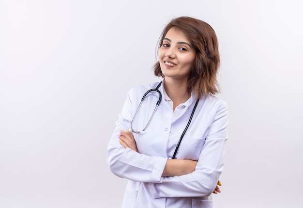 Foto gratuita médico joven en bata blanca con estetoscopio sonriendo confiado de pie con los brazos cruzados sobre la pared blanca