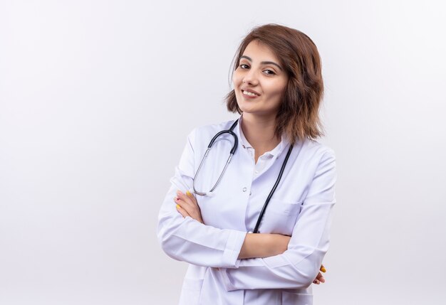 Médico joven en bata blanca con estetoscopio sonriendo confiado de pie con los brazos cruzados sobre la pared blanca