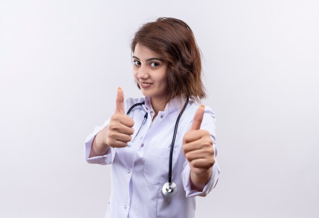 Médico joven en bata blanca con estetoscopio sonriendo mostrando los pulgares para arriba con ambas manos de pie sobre la pared blanca