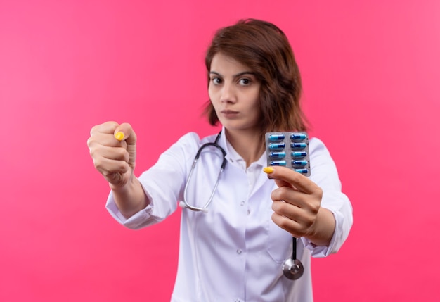 Médico joven en bata blanca con estetoscopio sosteniendo blister con pastillas mostrando el puño a la cámara con cara seria