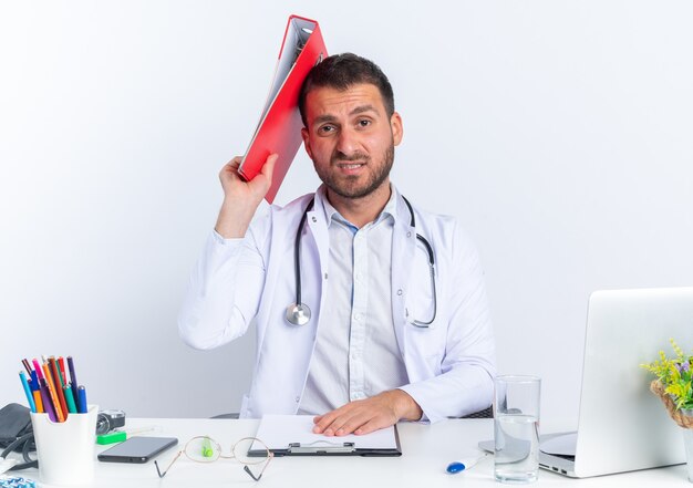 Médico joven en bata blanca y con estetoscopio sosteniendo la carpeta de la oficina en la cabeza mirando confundido sentado en la mesa con el portátil en blanco