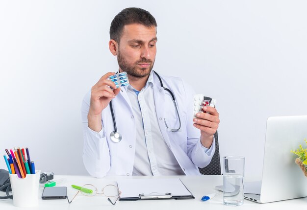 Médico joven en bata blanca y con estetoscopio sosteniendo diferentes pastillas mirándolos con cara seria sentado en la mesa con portátil sobre fondo blanco.