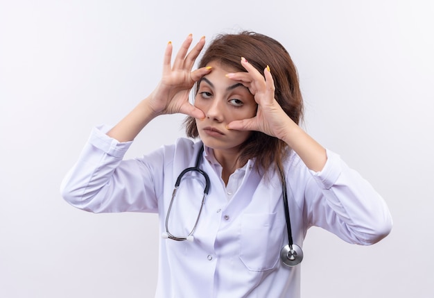 Médico joven en bata blanca con estetoscopio tratando de abrir los ojos con las manos sintiendo fatiga matutina de pie sobre la pared blanca