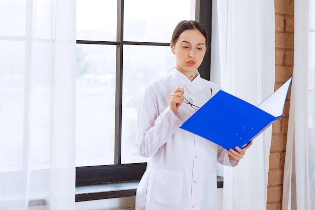 Médico joven en bata blanca leyendo sobre el próximo paciente cerca de la ventana.