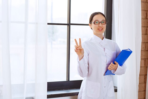 Médico joven en bata blanca posando con carpeta azul cerca de la ventana.