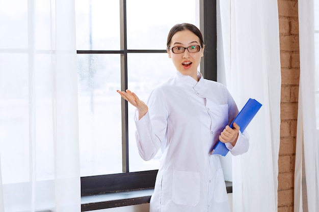 Médico joven en bata blanca posando con carpeta azul cerca de la ventana.