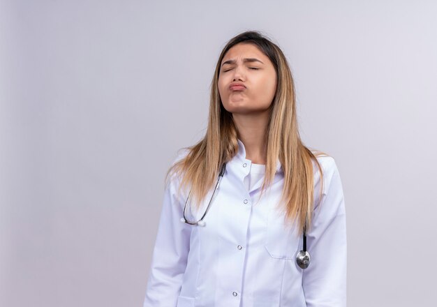 Médico joven y bella mujer molesta vistiendo bata blanca con estetoscopio soplando mejillas