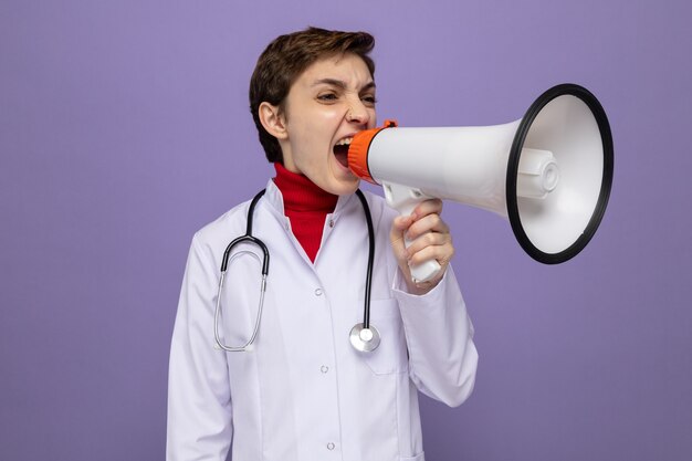 Médico joven emocional en bata blanca con estetoscopio alrededor del cuello gritando al megáfono con expresión agresiva de pie sobre la pared púrpura