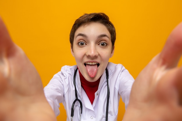 Médico joven feliz y divertido en bata blanca con estetoscopio alrededor del cuello tomando selfie sacando la lengua en naranja