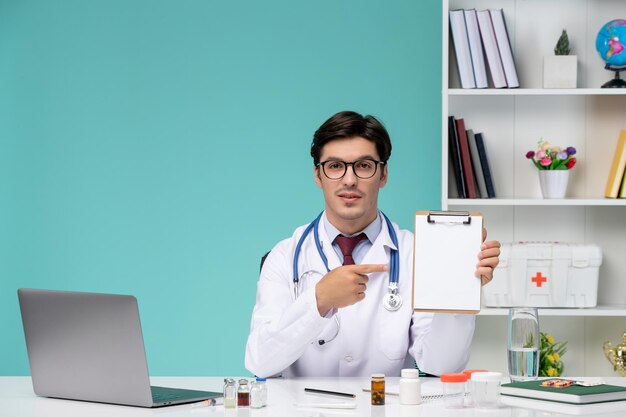 Médico joven inteligente en bata de laboratorio que trabaja de forma remota en una computadora con notas