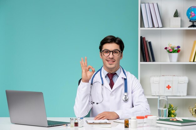 Médico joven inteligente en bata de laboratorio que trabaja de forma remota en una computadora que muestra el signo de ok