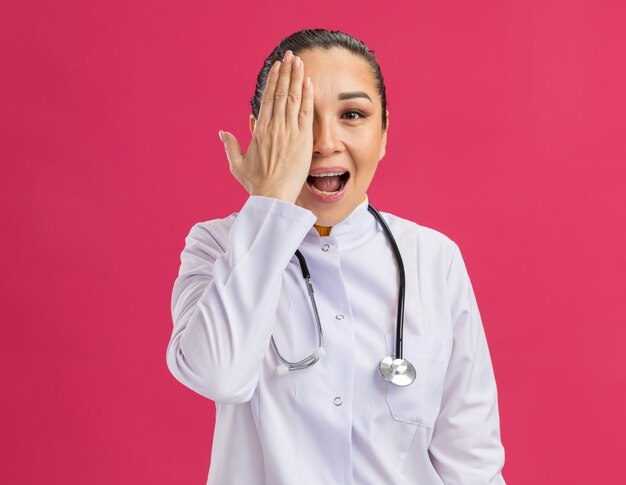 Médico joven sorprendido y feliz cubriendo un ojo con el brazo