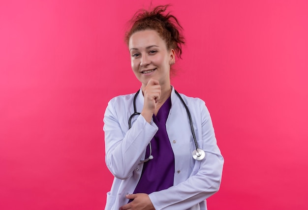 Médico joven vistiendo bata blanca con estetoscopio con la mano en la barbilla sonriendo de pie sobre la pared rosa