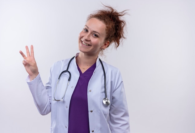 Médico joven vistiendo bata blanca con estetoscopio mostrando y apuntando hacia arriba con los dedos número tres sonriendo alegremente de pie sobre la pared blanca