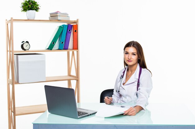 Médico con laptop sentada en el consultorio del médico hacer informe sonriendo aislado en la pared blanca