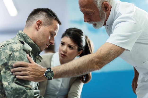 Médico maduro hablando con un soldado del ejército que vino con su esposa a una cita médica en el hospital El foco está en el soldado