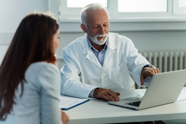 Médico maduro y su paciente hablando mientras leen el historial médico en una computadora en el consultorio del médico