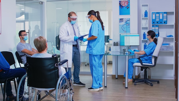 Médico con mascarilla contra covid19 discutiendo con la enfermera en la sala de espera del hospital. Mujer mayor discapacitada en silla de ruedas esperando examen. Asistente trabajando en la computadora de recepción.