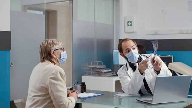 Médico masculino que analiza la exploración de radiografía con un paciente mayor en la visita de control. Médico explicando los resultados de rayos X y el diagnóstico de huesos a una mujer jubilada en un consultorio médico durante una pandemia.