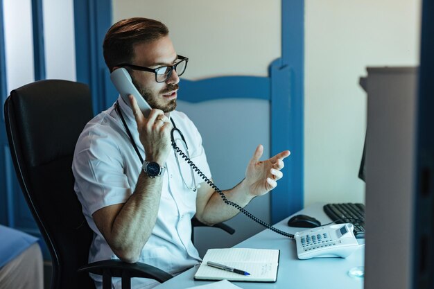 Médico masculino sentado en su oficina y comunicándose con alguien por teléfono