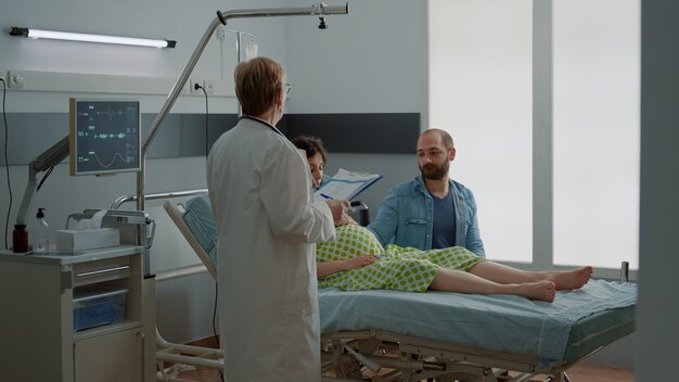 Médico de maternidad dando consejos a la mujer embarazada y el marido sentado en la sala del hospital. Joven pareja caucásica recibiendo asistencia médica para la paternidad de un especialista en parto en la clínica