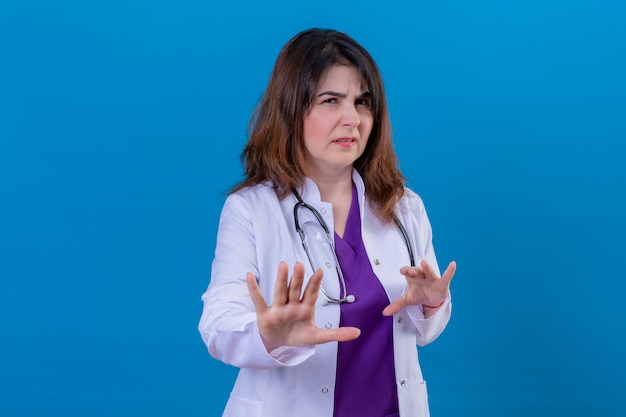 Médico de mediana edad con bata blanca y con estetoscopio levantando las palmas en gesto de parada y rechazo, expresión de disgusto sobre la pared azul aislada