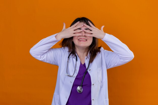 Médico de mediana edad con bata blanca y con estetoscopio sonriendo con las manos en la cara cubriendo los ojos sobre la pared naranja aislada