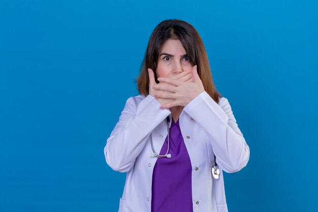 Médico de mediana edad con bata blanca y con estetoscopio sorprendido y sorprendido cubriendo la boca con las manos sobre la pared azul