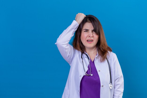 Médico de mediana edad con bata blanca y con estetoscopio se ve confundido y decepcionado al tocar su cabeza con la mano sobre la pared azul