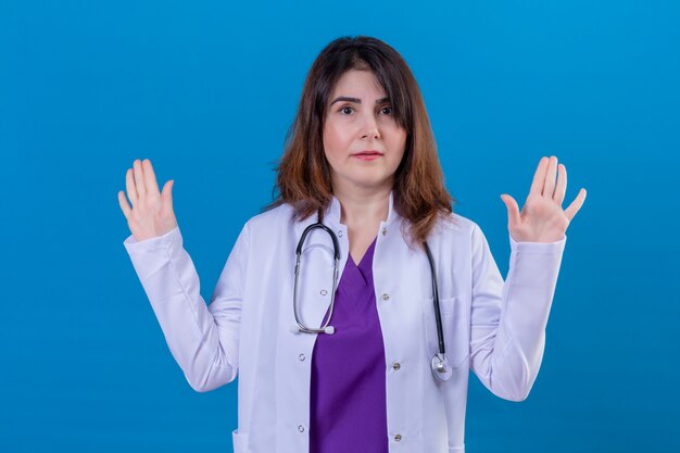 Médico de mediana edad vistiendo bata blanca y con estetoscopio levantando las manos en rendición mirando confundido sobre la pared azul