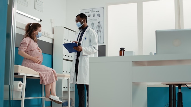 Médico y mujer embarazada con mascarilla facial que asisten a una visita de control para hacer una consulta médica en el consultorio. Médico general tomando notas en los archivos en la cita de examen con una persona embarazada.