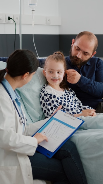 Médico mujer médico que controla los síntomas de la enfermedad escribiendo tratamiento con antibióticos durante la consulta de recuperación en la sala del hospital. Niño hospitalizado con tubo nasal recuperándose después de una cirugía respiratoria