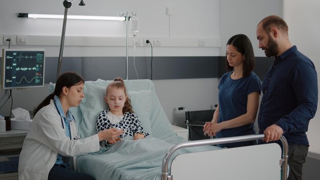 Médico de la mujer practicante que pone el oxímetro médico en el dedo del paciente del niño pequeño que monitorea el pulso del latido del corazón durante la consulta de recuperación. Enfermera negra comprobando la bolsa de goteo intravenosa trabajando en la sala del hospital