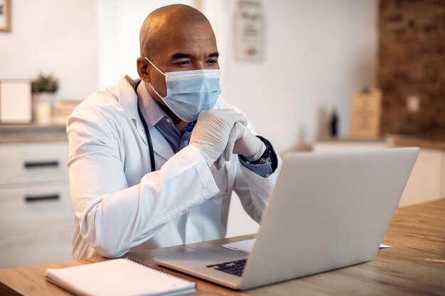 Médico negro con mascarilla protectora trabajando en una computadora en su oficina