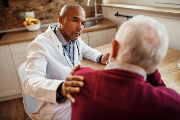 Médico negro reconfortante anciano mientras habla con él durante la visita a casa