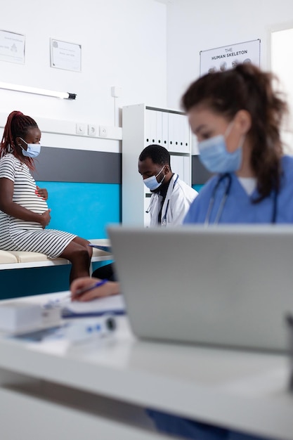 Médico y paciente con mascarillas hablando sobre el embarazo en el chequeo médico en la oficina. Mujer afroamericana esperando un hijo y haciendo consultas médicas con el médico.