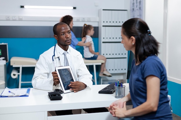 Médico pediatra afroamericano con tableta con radiografía de niña en pantalla explicando el tratamiento médico a la madre del paciente durante la cita en el consultorio del hospital. Servicio de medicina