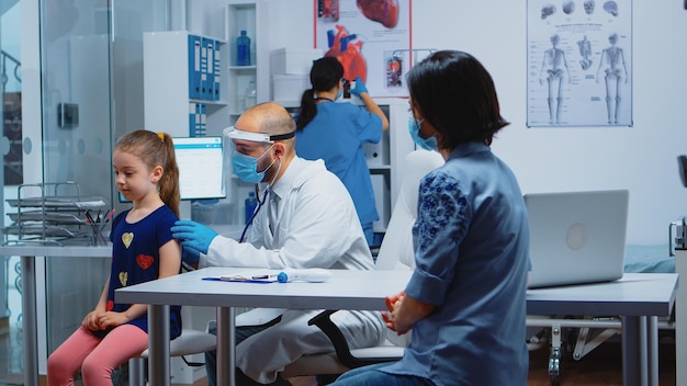 Médico pediatra con máscara de protección y estetoscopio escuchando aliento de niña. Médico especialista en medicina que brinda servicios de atención médica, consulta, tratamiento durante el covid-19 en el hospital