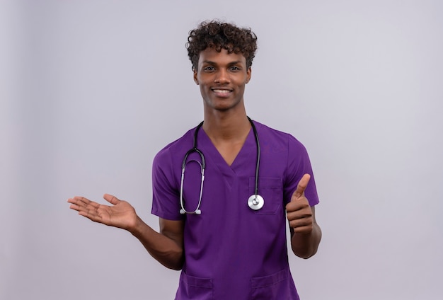 Un médico de piel oscura guapo joven complacido con cabello rizado vistiendo uniforme violeta con estetoscopio con pulgares hacia arriba