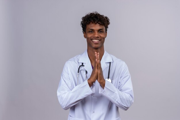 Un médico de piel oscura guapo joven complacido con pelo rizado vistiendo bata blanca con estetoscopio mostrando gesto de agradecimiento con las manos