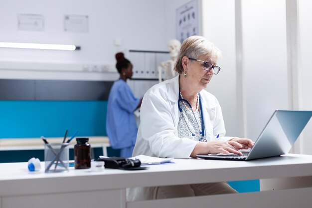 Médico principal que usa una computadora portátil en el gabinete para buscar tratamiento de atención médica. Médico general mirando la pantalla de la computadora para trabajar en medicamentos recetados contra el diagnóstico de enfermedades.