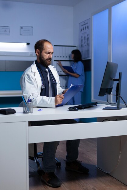 Médico con problemas trabajando con monitores y papeles sanitarios para consulta y examen. Médico que usa la computadora para obtener información médica y análisis en horas extraordinarias. Practicante de la salud