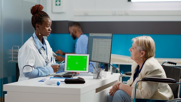 Médico que sostiene una tableta con pantalla verde horizontal en la cita con una mujer en silla de ruedas. Uso de pantalla de clave cromática con espacio de copia aislado y fondo de maqueta en blanco. Disparo de trípode.