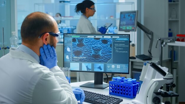 Médico químico preocupado trabajando en pc examinando la evolución de la vacuna en un laboratorio científico moderno equipado